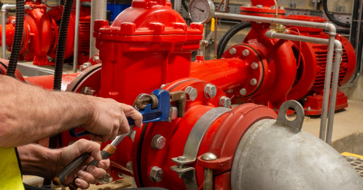 A close-up view shows a person wearing a yellow safety vest using a blue wrench to tighten a bolt on a large, red pipe.