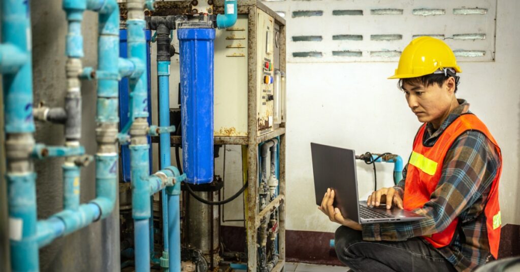 A man wearing an orange safety vest squats in front of a system of blue pipes and types on a laptop.