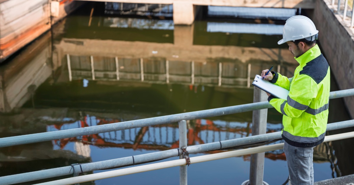 A man wearing a neon-yellow jacket and a white hard hat is writing on a clipboard standing next to water treatment pool.