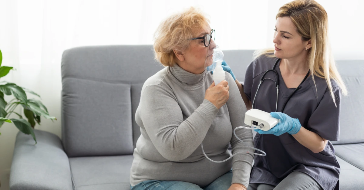 A medical professional is holding a respiratory device for a senior female patient. They're sitting on a gray couch.