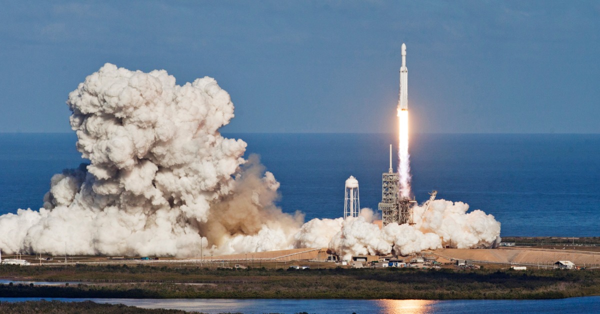 A white satellite is being launched next to a large body of water. A large plume of smoke forms on the ground.