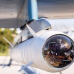 A close-up view shows the end of a missile that's attached to the wing of an aircraft. The end is clear.