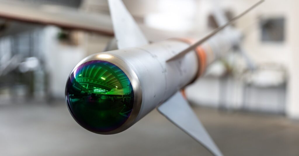 A close-up view shows the tip of a missile with a reflective covering. Three metal wings are behind the tip.