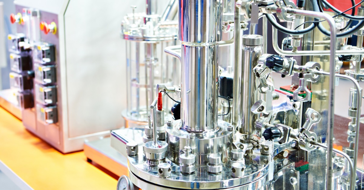 A metal bioreactor sits on a table with a yellow counter. It sits next to other biotech equipment.