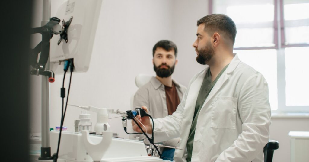 A male medical professional sits on a black stool and shows a patient an endoscopic tool in an exam room.