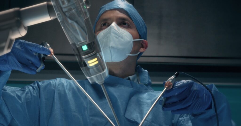 A surgeon wearing a white mask, blue latex gloves, and a blue surgical cap is operating a robotic surgical tool.