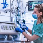 A female medical professional wearing a face mask and teal scrubs is looking at a medical ventilator in a hospital setting.