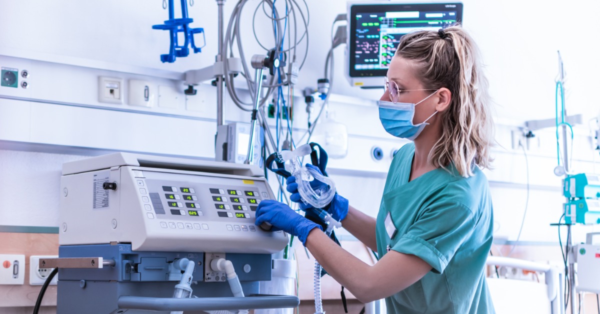 A female medical professional wearing a face mask and teal scrubs is looking at a medical ventilator in a hospital setting.