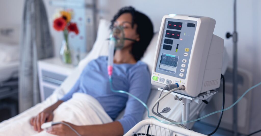A female patient lies in a hospital bed wearing an oxygen mask that's hooked up to a ventilator monitor.