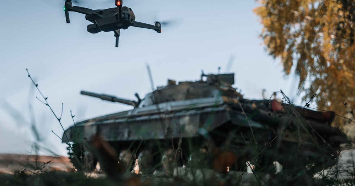 A drone with four propellers is flying in the air in a grassy area. A large military tank is in the background.